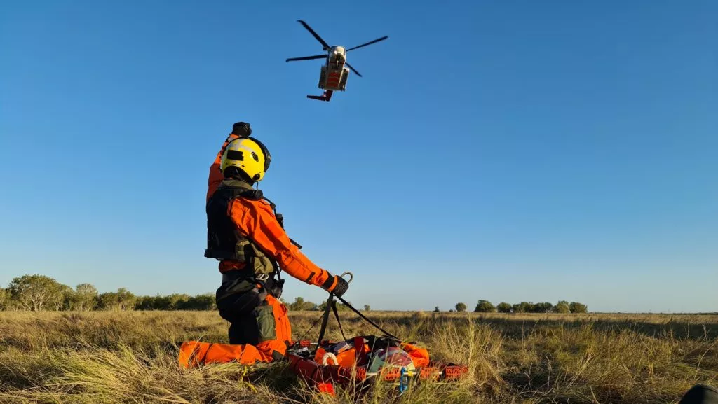 Search and rescue (SAR) crewmember landed in field with PHI red and yellow SAR aircraft flying overhead