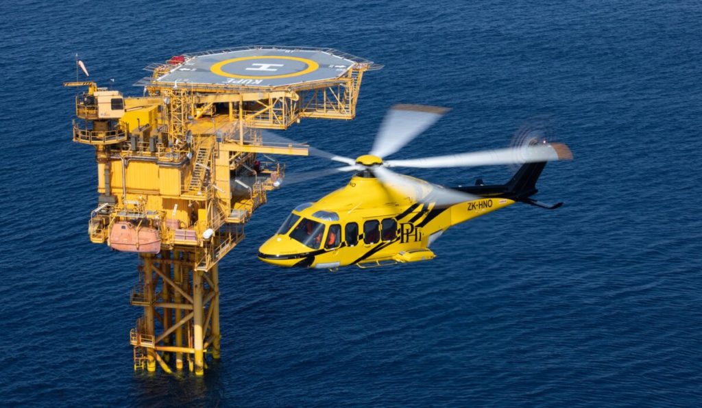 Yellow and black painted AW139 with PHI logo flying offshore over water with oil rig platform in background
