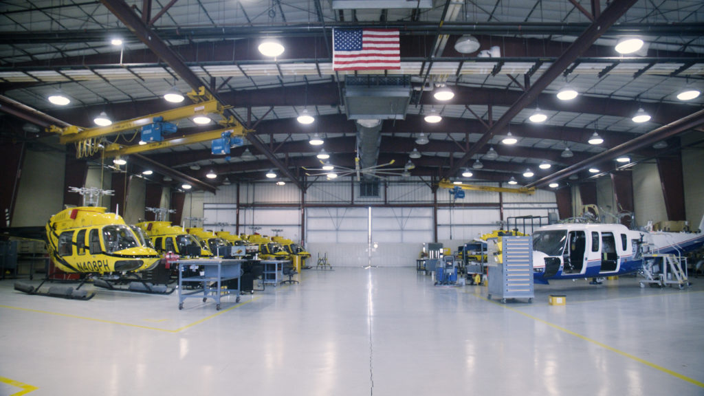 Helicopter hangar with several yellow and black helicopters lined up on left-hand side and white and blue helicopter on right-hand side