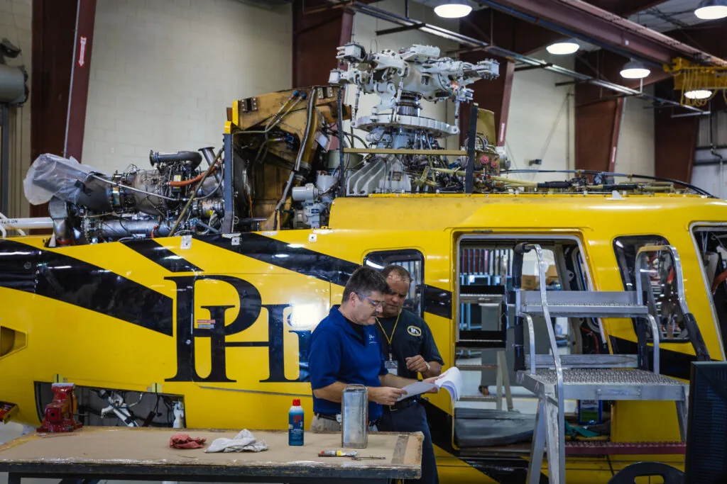 Two PHI mechanics look at papers in front of PHI yellow and black helicopter in hangar
